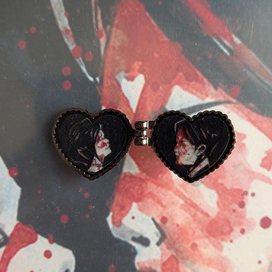 A pair of heart-shaped black enamel pins with a small silver clasp, featuring three cheers for sweet revenge, my chemical romance character artwork, displayed on a red and black illustrated background.